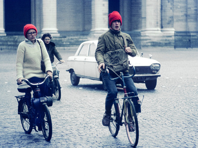 A woman and man are shown, with the woman riding a Solex and the man on a bicycle. Both are wearing red woolly hats, and a white car is in the background.