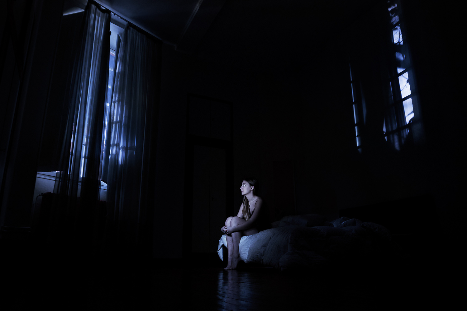 Une jeune fille est assise sur un lit. Elle est dans la pénombre, seule la lumière de la lune éclaire la pièce. Photographie de Johanna Senpau à Toulouse.