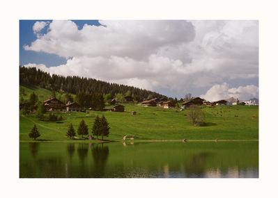 Fine Art print edition of 7 signed & numbered. 50x70 or 30x40 cm on Hahnemühle Bright White paper. Olivier Montay photographer. White clouds overhang chalets spread out on a grassy slope. France, lac des Confins. Lake. Film photography