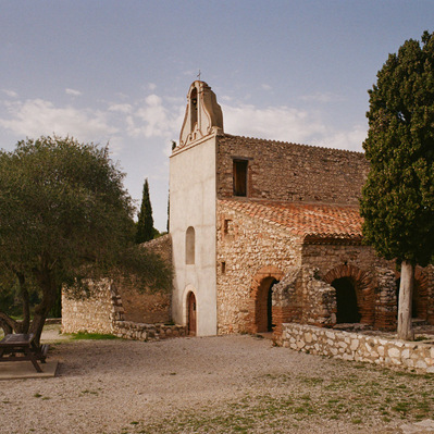 The Saint-Vincent chapel, with rough stone walls and an ocher tiled roof. A bushy tree in front of the arcades. A bench and a wooden table placed on the gravel, in front of the gray facade. France, Occitanie, Pyrénées-Orientales, Estagel. 2022