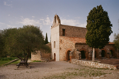 The Saint-Vincent chapel, with rough stone walls and an ocher tiled roof. A bushy tree in front of the arcades. A bench and a wooden table placed on the gravel, in front of the gray facade. France, Occitanie, Pyrénées-Orientales, Estagel. 2022