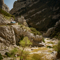 A Volkswagen combi is stopped under a hot sun on a road on the side of a cliff. In the middle of these rocky gorges flows weakly the Agly river. France, Occitanie, Pyrenees-Orientales, Gorges de Galamus, April 2022.