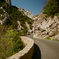 A road bordered by a low stone wall winds between the light stone cliffs strewn with vegetation in the Galamus gorges. Central perspective.
Une route bordée d'un muret de pierres claires serpente entre les falaises. Point de fuite central.