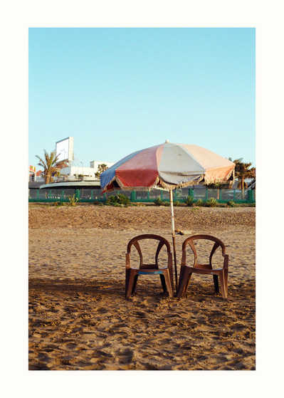 Fine Art print edition of 7 signed & numbered. 50x70 or 30x40 cm on Hahnemühle Bright White paper. Olivier Montay photographer. Two brown plastic armchairs, discolored parasol, beach sand. Aïn Diab. Morocco. Film photography