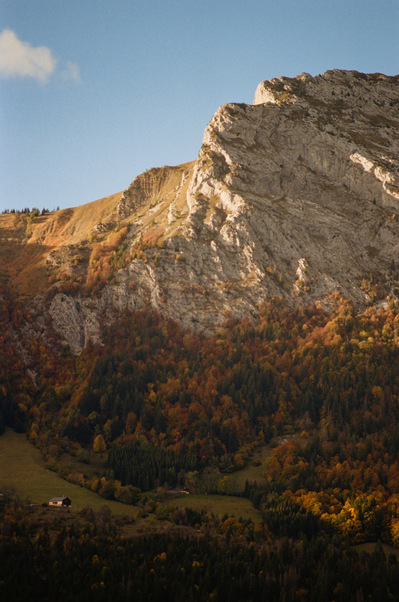 Rocky summit of a mountain lighted by sunrise. Its slopes and autumn vegetation remain in shadow. At the bottom of the slope, in a grassy clearing, a chalet is still plunged into darkness. France, Auvergne-Rhône-Alpes, Haute-Savoie, Thônes, 2021