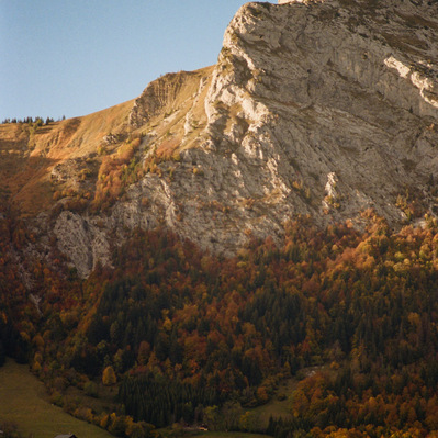 Rocky summit of a mountain lighted by sunrise. Its slopes and autumn vegetation remain in shadow. At the bottom of the slope, in a grassy clearing, a chalet is still plunged into darkness. France, Auvergne-Rhône-Alpes, Haute-Savoie, Thônes, 2021