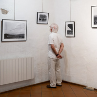 A man dressed in white, in the corner of white walls, in front of black and white photos by photographer Corentin Schimel. "Regards croisés : dans les traces de François Arago" exhibition. 2022 September, France, Occitanie, Pyrénées Orientales, Estagel