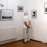 A man dressed in white, in the corner of white walls, in front of black and white photos by photographer Corentin Schimel. "Regards croisés : dans les traces de François Arago" exhibition. 2022 September, France, Occitanie, Pyrénées Orientales, Estagel