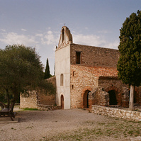 The Saint-Vincent chapel, with rough stone walls and an ocher tiled roof. A bushy tree in front of the arcades. A bench and a wooden table placed on the gravel, in front of the gray facade. France, Occitanie, Pyrénées-Orientales, Estagel. Avril 2022
