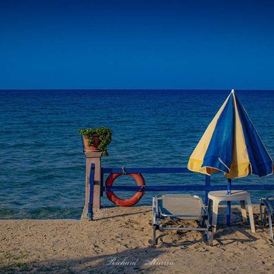 A high quality print available to buy of a lonely closed sunshade and beach loungers waiting for the return of the tourists in Greece.