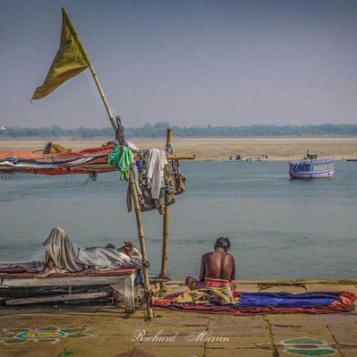 A high quality print available to buy of By the Ghat, Varanasi, India, showing a man seated and another reclining by the Ganges.