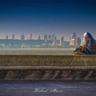 A high quality print available to buy of an elderly lady sitting on the riverbank at Mumbai.