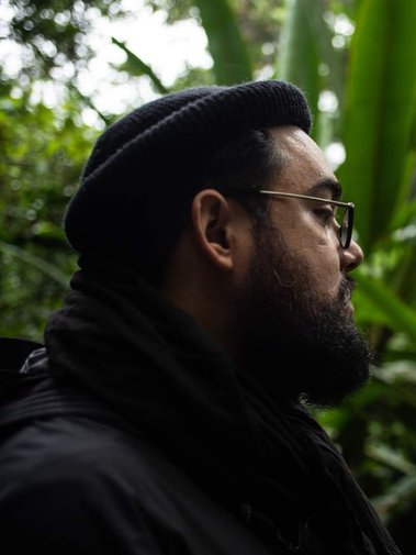 Ash Ponders wears an inscrutable beanie in a jungle, they look left.