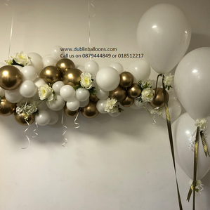 A gold and white garland with flower accessories and three white large balloons for celebrations.
