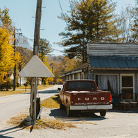 A vintage ford truck sits outside an abandoned house in the midday sun