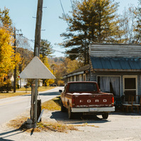 A vintage ford truck sits outside an abandoned house in the midday sun