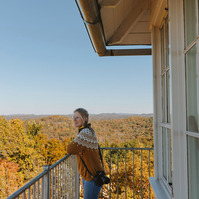 Blonde woman stands on balcony of old fire tower looking over yellow and orange trees