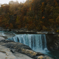 Long exposure of a waterfall cascading down, with smooth rocks in the foreground and yellow and orange trees behind.