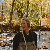A blonde woman smiling as she looks over to her right, holding a fishing rod standing in the river
