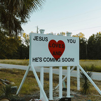 A white painted wooden sign sits on a road side saying: Jesus loves you, he's coming soon. Rubbish sits at the base of the sign.