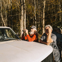 Two men lean on the bonnet of an old white truck, one with binoculars.