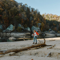 A blonde woman in a red and white t-shirt balances on a fallen tree trunk. Evening light shines on the beach behind her.