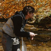 A young dark haired man leaning forward as he flicks his fishing rod, yellow and orange leaves shine in the background