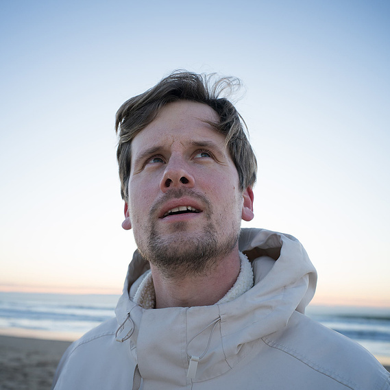 Portrait en extérieur d’un photographe au bord de l’océan au coucher du soleil, capturant une expression contemplative et authentique, symbolisant la curiosité et l’immersion dans les environnements variés.
