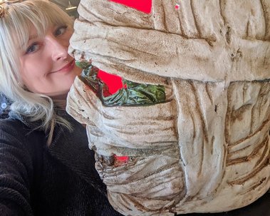 Jay is a white woman with blonde hair. She poses with a Halloween decoration. It is a mummy head which illuminates uncanny red light from within.