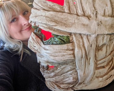 Jay is a white woman with blonde hair. She poses with a Halloween decoration. It is a mummy head which illuminates uncanny red light from within.