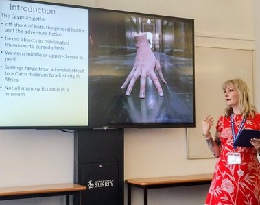 Jay is a white woman with blonde hair. She presents at a conference in a red floral summer dress. On the PowerPoint located on the television screen behind her is a slide with a picture of Thing from Wednesday. The slide reads:
Introduction