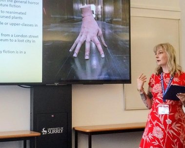 Jay is a white woman with blonde hair and a red dress. She presents at a conference and gesticulates mid-sentence. On the TV screen behind her is a PowerPoint showing a disembodied hand.