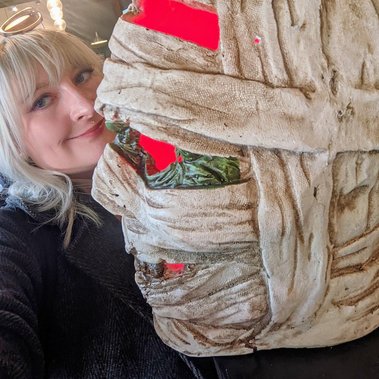 Jay is a white woman with blonde hair. She poses with a light-up mummy head which may be a Halloween decoration.  