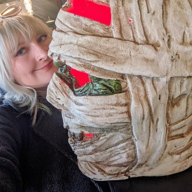 Jay is a white woman with blonde hair. She poses with a light-up mummy head which may be a Halloween decoration.  