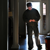 Jay, backlit, buttons his shirt in the entryway of his home.