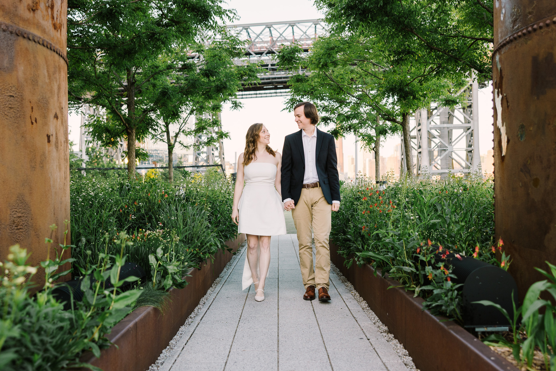 Brooklyn domino park engagement session, beautiful couple walking hand in hand, green vegetation, leading lines and smiles. captured by mc3 photo, documentary nyc engagement photographer 