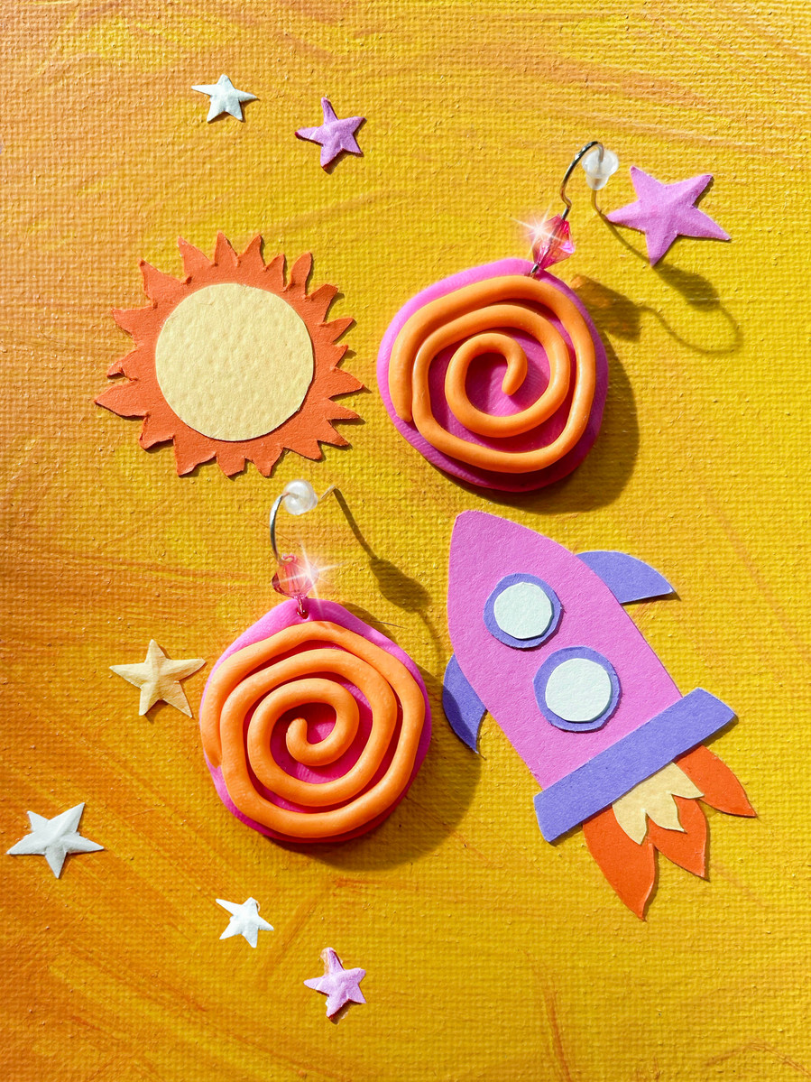 Handmade polymer clay earrings in pink and orange colour with a spiral design. Dangle earrings photographed on a yellow and orange background  consisting of paper craft cut-outs. Paper sun paper rockets and paper stars. 