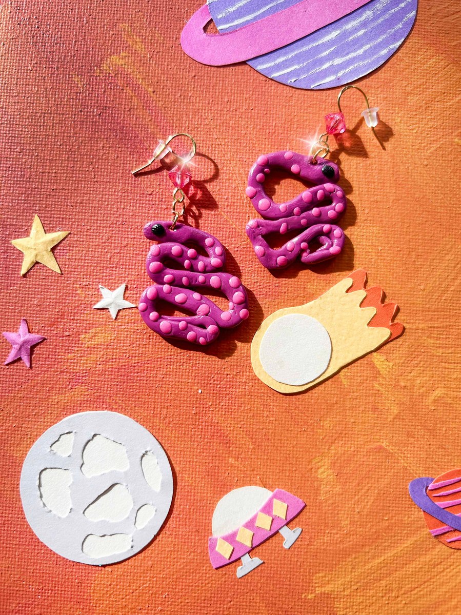 Purple and pink polymer clay earrings for kids. Handmade and lightweight earrings great for kids parties and return favours. Polka dotted snake children's jewellery. Handmade orange, purple and pink paper art background.