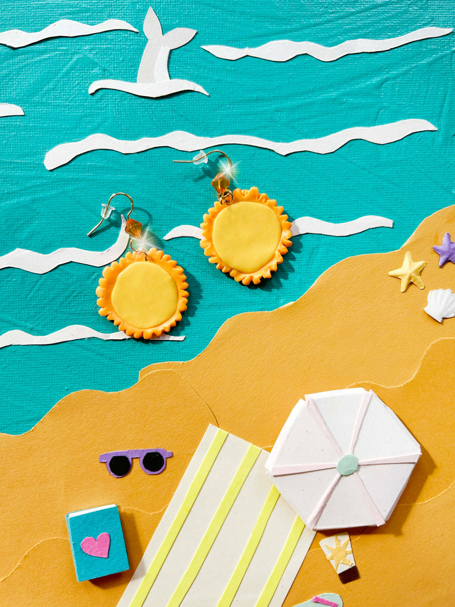 Handmade polymer clay yellow sun shaped earrings photographed on a blue and yellow beach scene background. The background has handmade paper cutout elements such as a paper umbrella, paper dolphin, paper sunglasses, paper books and paper sea shells. 