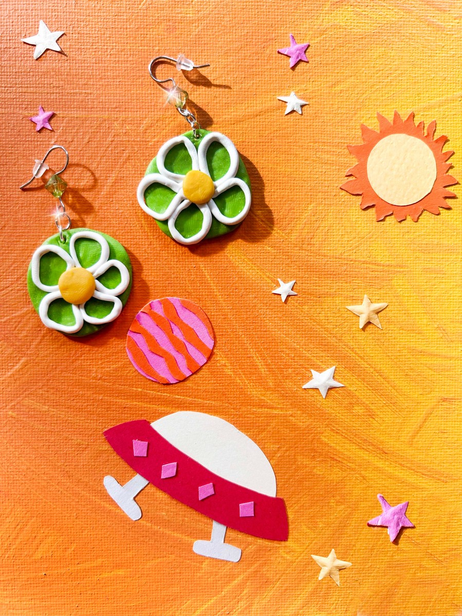 Handmade flower shaped green and yellow polymer clay dangle earrings. Photographed on a colourful orange background that has paper cutout elements of outer space scene. Paper sun, paper stars, paper planets, paper ufo cutout. Fun paper cutouts for kids.