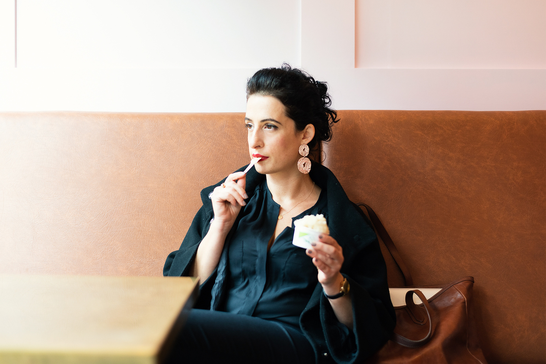 Portrait of woman elegantly dressed on a black shirt, sitting on a brown leather couch and a brass table, eating an ice-cream while looking out of the window with a confident gaze.