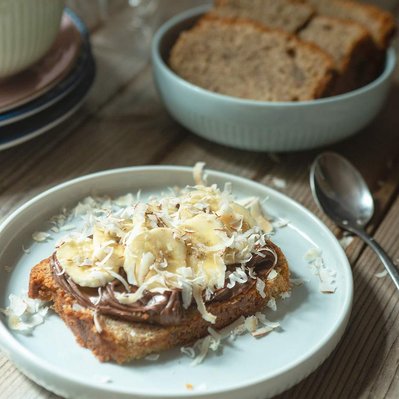 slice of banana bread with a spread of chocolate spread, sliced bananas and roasted coconut gratings on a plate with more banana bread slices in the background and coffee glass mugs