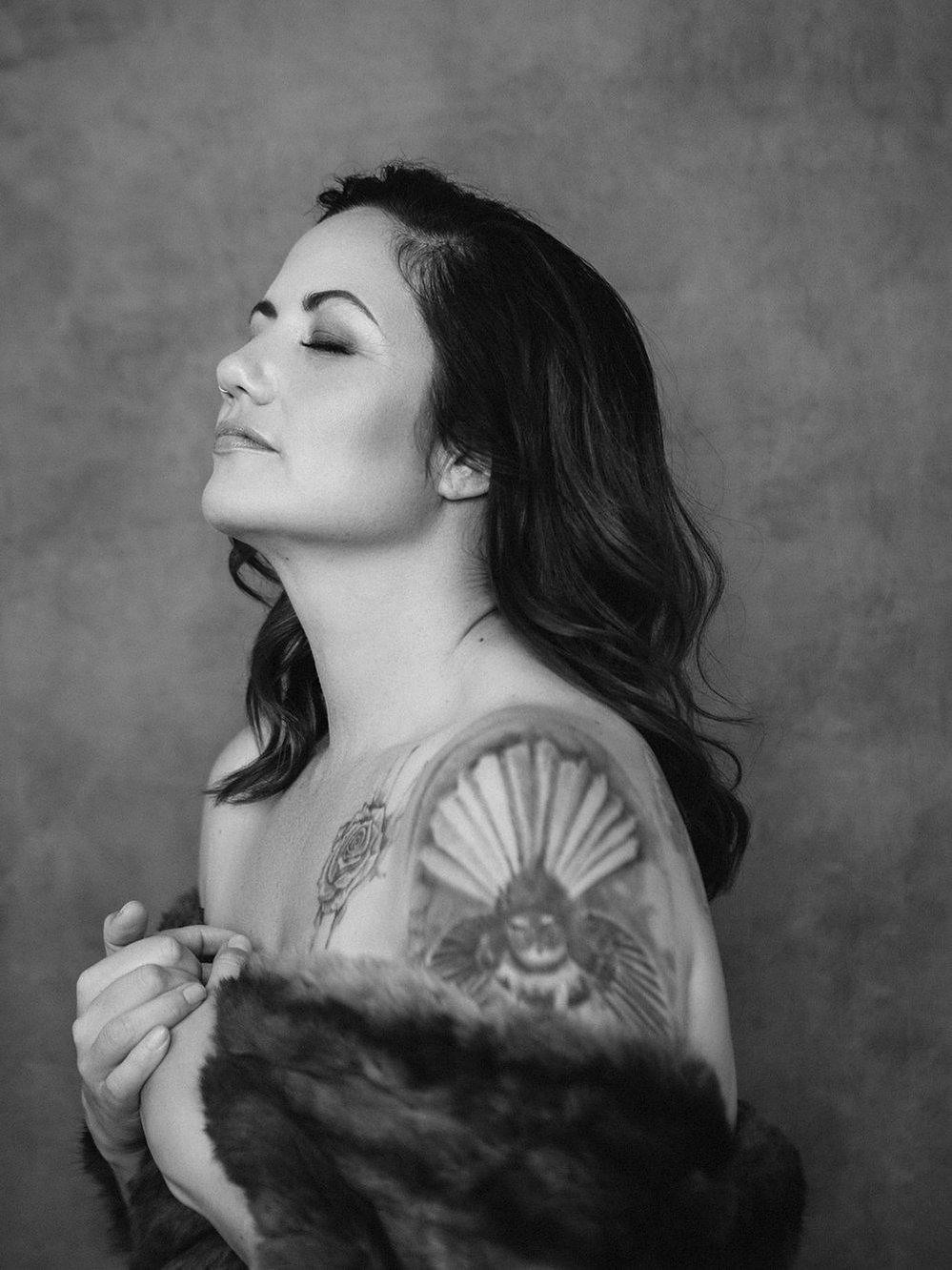 A glamourous young brunette woman is wrapped in a fur, her shoulders visible and displaying a large fantail tattoo