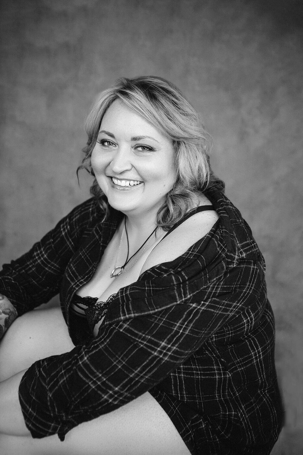 A curvy blonde woman with tattoos wears a flannel shirt and smiles