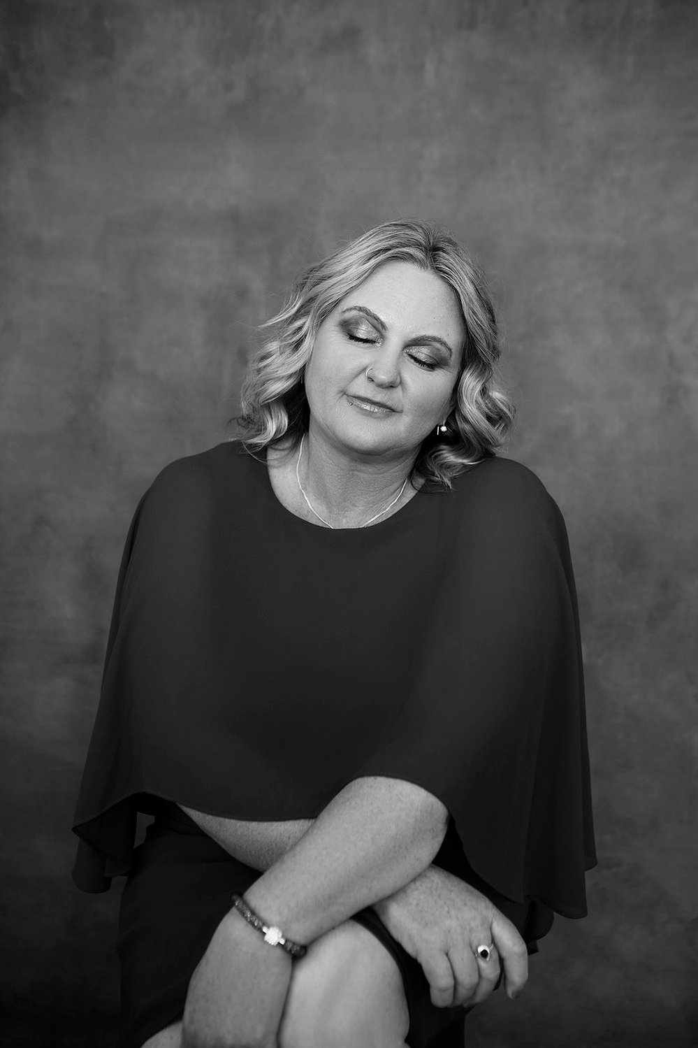 A curvaceous woman with a blonde bob sits with eyes closed and her hands crossed
