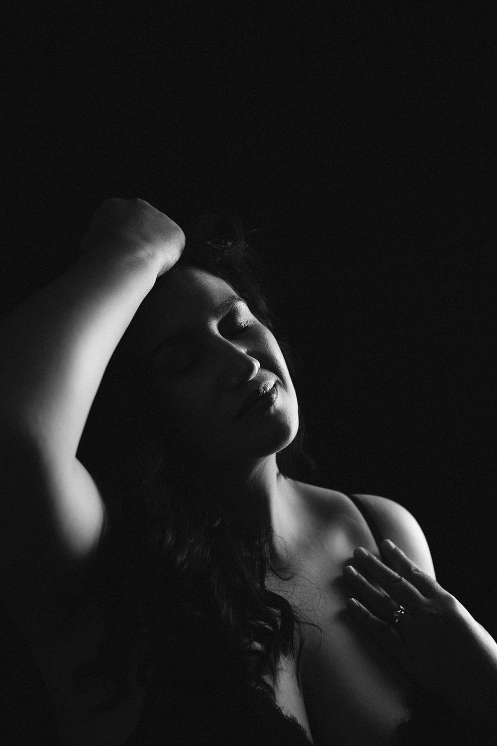 A stunning brunette woman is illuminated from one side, she runs one hand through her hair and the other traces her collarbone
