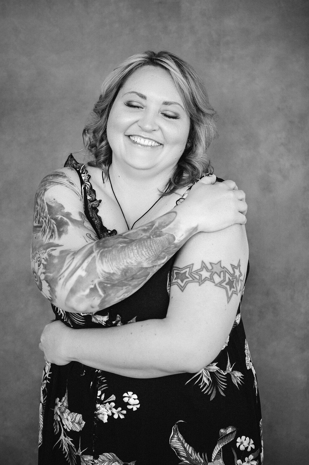 A curvy blonde woman with tattoos wears a floral dress as she embraces herself