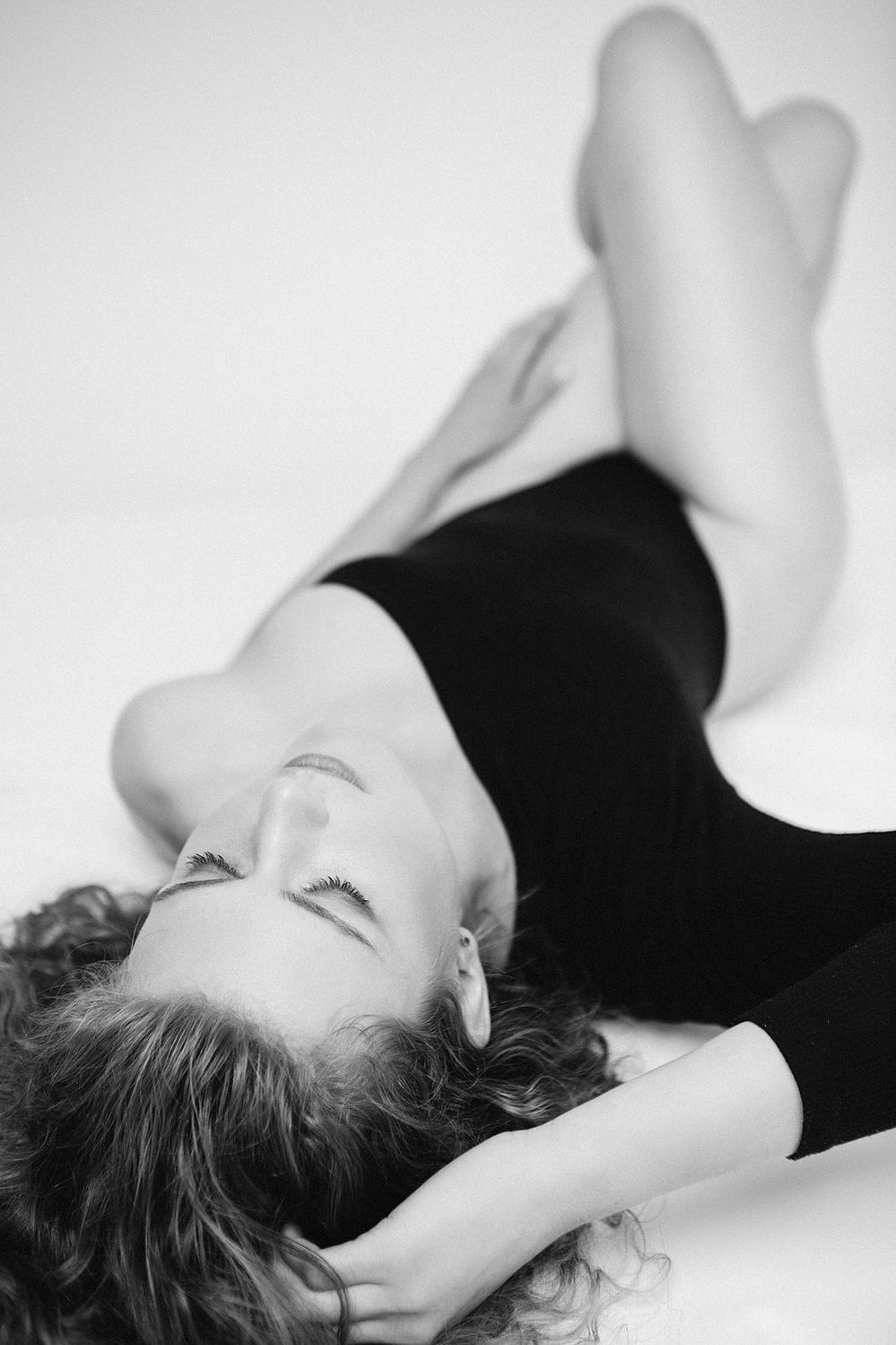 A young woman with blonde naturally curly hair wears an asymmetrical bodysuit, she reclines on a bed and has a hand in her hair