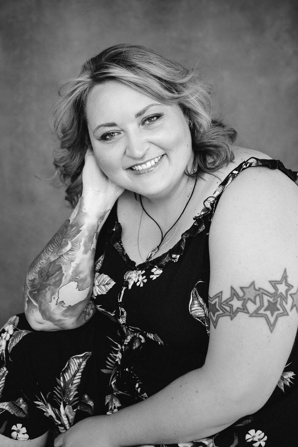 A curvy blonde woman with tattoos smiles at the camera with her head resting on one of her hands