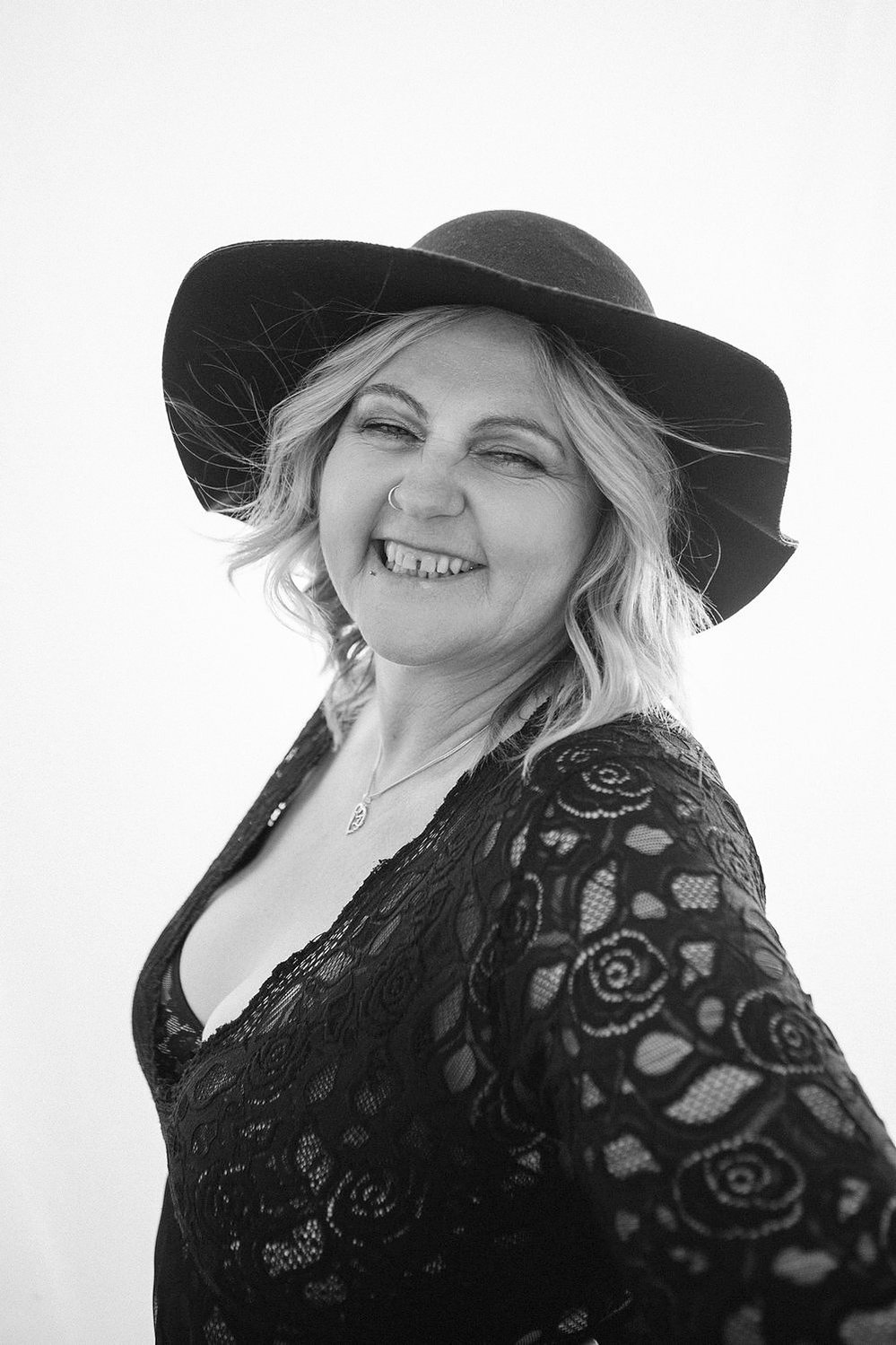 A curvaceous woman with a blonde bob wears a wide brimmed hat and lace bodysuit
